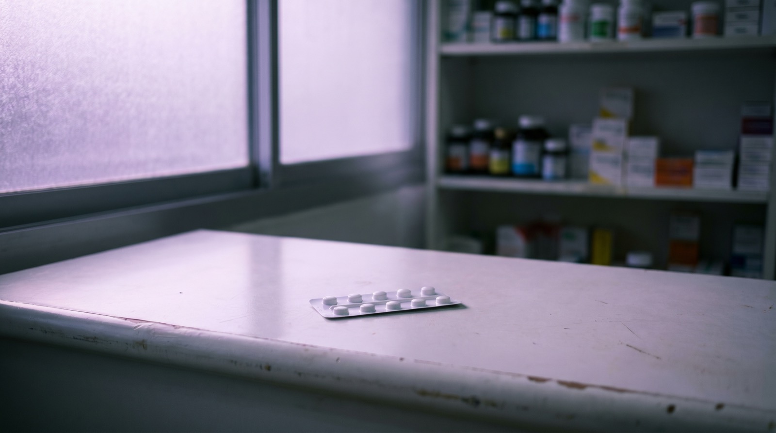 A quiet pharmacy counter at dawn: a single closed white blister pack of unlabeled tablets resting flat on a white surface, soft violet ambient light from frosted glass window, no people, no text visible
