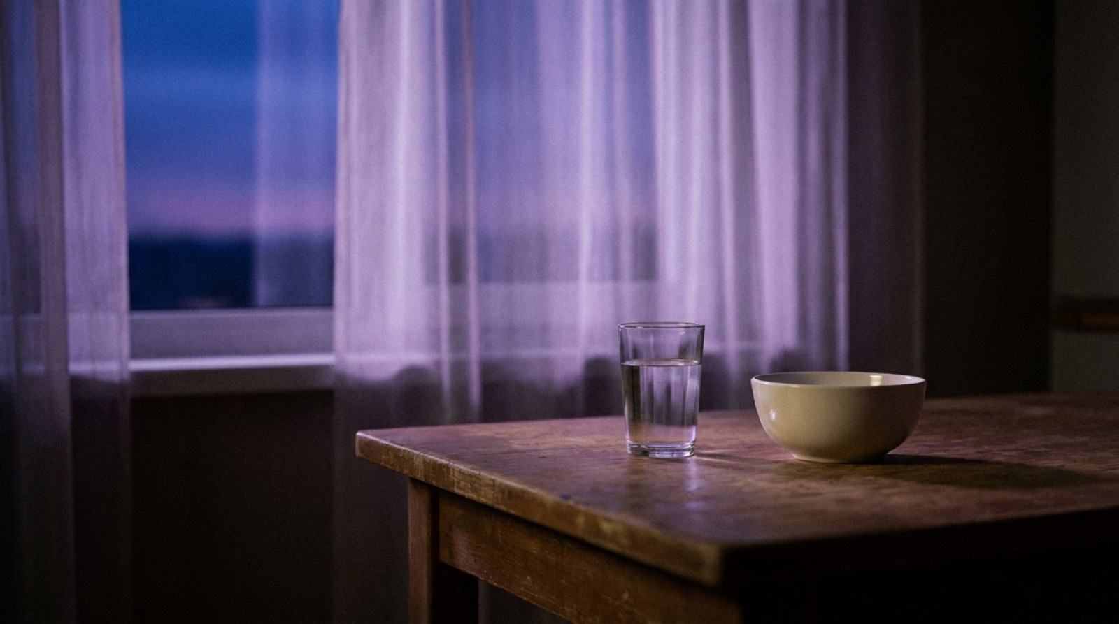A quiet kitchen table near a window at dusk, a single glass of water and a plain bowl lit by soft violet daylight — calm, unhurried, no people.