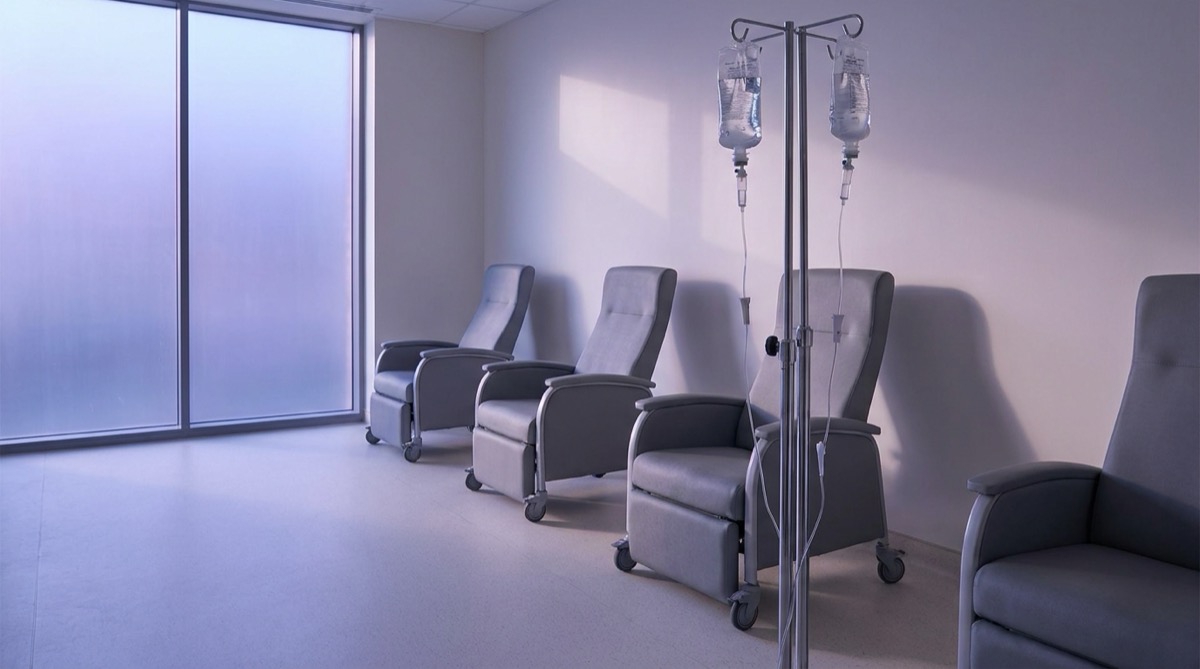 Empty hospital infusion suite at dawn: a tall chrome IV drip stand with two clear infusion bags in the foreground, reclining clinical chairs arranged along a pale wall, soft violet ambient light filtering from a frosted side window, no people, no visible text or labels on any surface, photographic realism, depopulated interior still-life