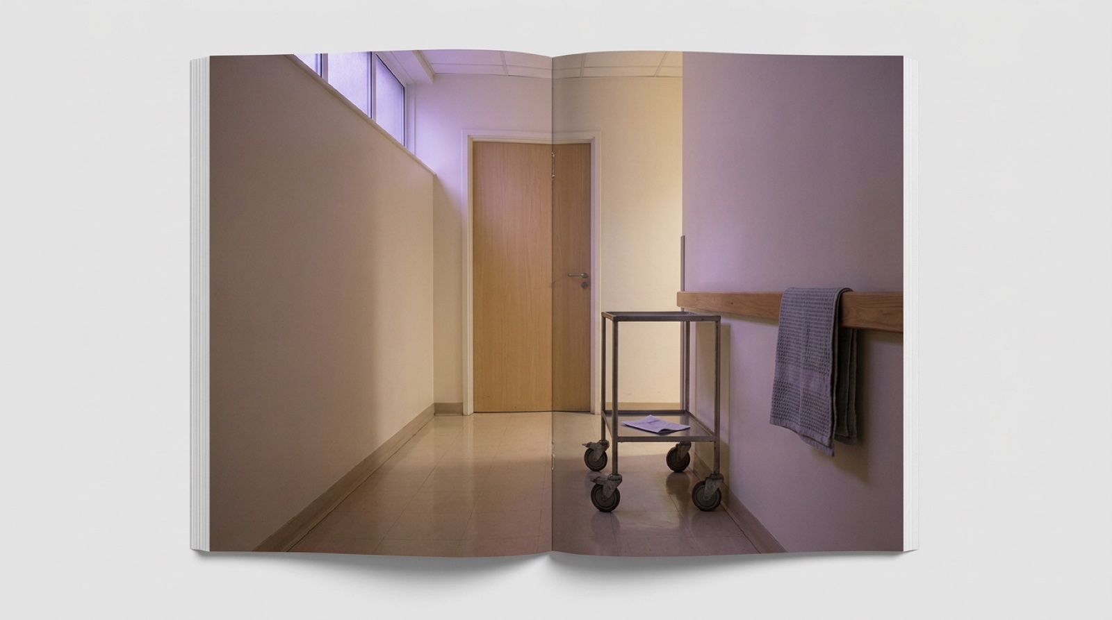 A quiet hospital corridor at dawn: a polished pale-tile floor receding past a closed light-wood ward door beside a small empty wheeled equipment cart, soft violet ambient light from a high clerestory window, no people, no signage visible.
