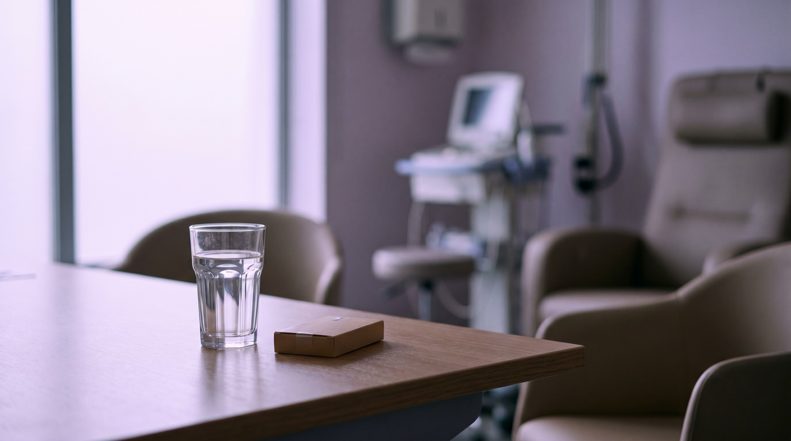 A quiet, empty consultation desk with a glass of water and a small sealed medication box, soft violet ambient light from a side window, no people, no text, calm clinical setting